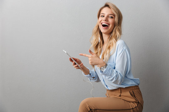 Photo of excited blond businesswoman with long curly hair rejoicing while listening to music with smartphone and earphones
