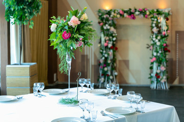 Beautiful table setting with crockery and flower arrangement in a vase on a high stem for a party, wedding reception or other festive event. Glassware and cutlery for catered event dinner