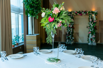 Beautiful table setting with crockery and flower arrangement in a vase on a high stem for a party, wedding reception or other festive event. Glassware and cutlery for catered event dinner
