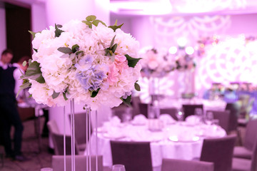 Beautiful table setting with crockery and white flower arrangements in a vase on a high stem for a party, wedding reception or other festive event. Glassware and cutlery for catered event dinner