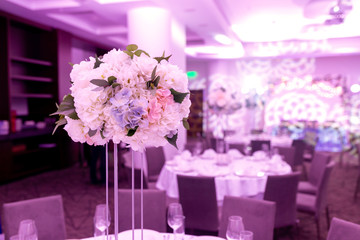 Beautiful table setting with crockery and white flower arrangements in a vase on a high stem for a party, wedding reception or other festive event. Glassware and cutlery for catered event dinner