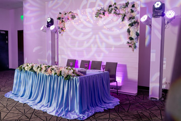 Wedding banquet in blue and white tones. Dinner table for newlyweds decorated with greenery and long cloth. A long flower arrangement on the table and a floral decor on a white wooden wall