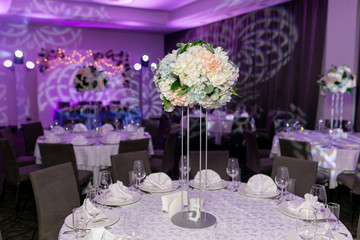 Beautiful table setting with crockery and white flower arrangements in a vase on a high stem for a party, wedding reception or other festive event. Glassware and cutlery for catered event dinner