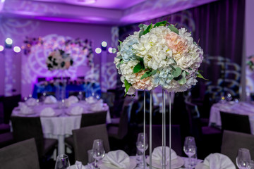 Beautiful table setting with crockery and white flower arrangements in a vase on a high stem for a party, wedding reception or other festive event. Glassware and cutlery for catered event dinner