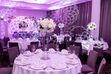 Beautiful table setting with crockery and white flower arrangements in a vase on a high stem for a party, wedding reception or other festive event. Glassware and cutlery for catered event dinner