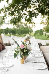 Wedding table setting decorated with fresh flowers in a brass vase. Wedding floristry. Banquet table for guests outdoors with a view of green nature. Bouquet with roses, eustoma and eucalyptus leaves
