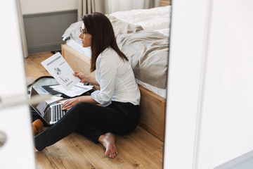 Portrait of elegant adult businesswoman working with paper documents and laptop while sitting on floor near luggage in apartment