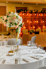 Beautiful table setting with crockery and white flower arrangements in a vase on a high stem for a party, wedding reception or other festive event. Glassware and cutlery for catered event dinner