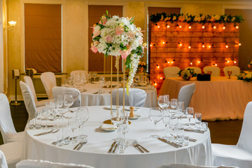 Beautiful table setting with crockery and white flower arrangements in a vase on a high stem for a party, wedding reception or other festive event. Glassware and cutlery for catered event dinner