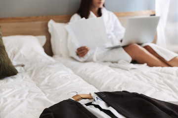 Blurred photo of elegant adult businesswoman working with paper documents and laptop while lying on bed in apartment