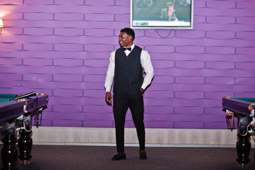 Young handsome african man wearing white shirt, black vest and bow tie standing against pool billiard tables.