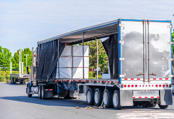 Big rig semi truck with open covered semi trailer unloading commercial cargo on warehouse parking lot