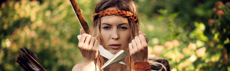 Beautiful girl archer with long blond hair with bow and arrowson a back holding two steel knifes...