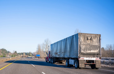 Day cab red big rig semi truck transporting commercial cargo in black covered semi trailer running on the straight wide highway