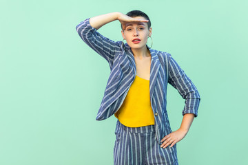 Portrait of attentive handsome beautiful short hair young stylish woman in casual striped suit standing with hand on forhead looking seriously. indoor studio shot isolated on light green background.