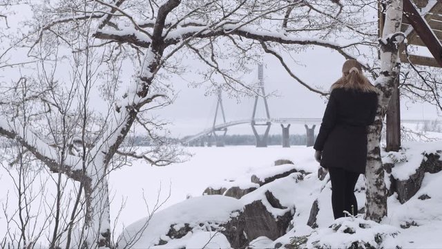 Wide Shot of a blonde woman waiting near a tree someone while looking at Replot Bridge in the distance. Vaasa (Finland) 
Shot on Red Monster 8K. In 4K