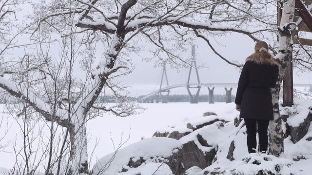 Wide Shot of a blonde woman waiting near a tree someone while looking at Replot Bridge in the distance. Vaasa (Finland) 
Shot on Red Monster 8K. In 4K
