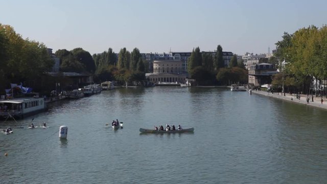 Paris, France - April 5, 2019: Parisian basin with people canoeing kayak during the summer with a beautiful blue sky. Pan to left. Lot of water, Seine. Static shot. Rotonde de La Villette background.