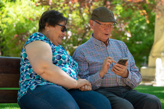 Pareja de gente mayor (abuelos) mostr&aacute;ndose mensajes, im&aacute;genes y v&iacute;deos enviados por mensajer&iacute;a instant&aacute;nea mientras se r&iacute;en y est&aacute;n sentados en un parque.