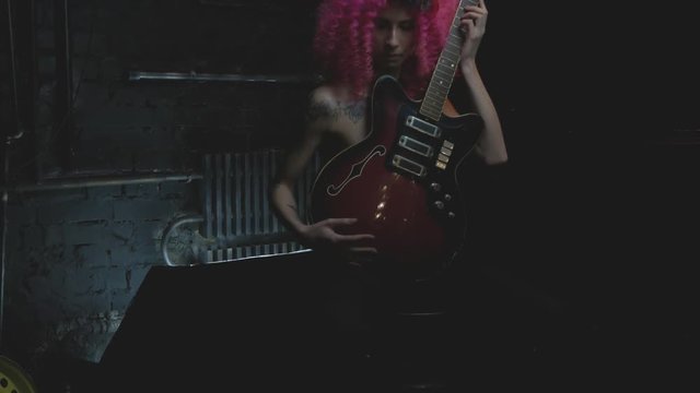 curly pink haired sexual girl posing with electric guitar in studio dark room. Rough rock metal style. Flower woven in hair. 