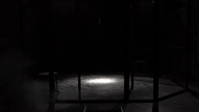 White smoke Projection in a metal grilles cage, circular MMA boxing ring, in a dark studio with a Spotlight or light shower. Show with Vintage microphone. Tracking in. Smoke coming from the ground.