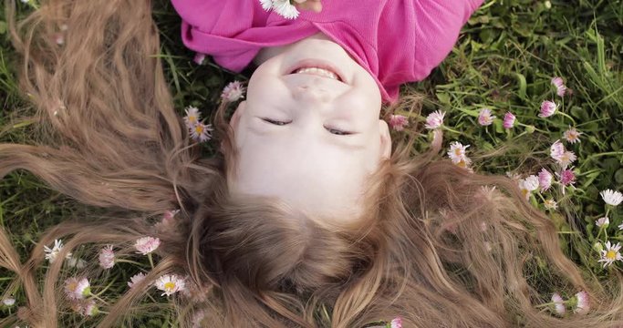 Close-up smiling little cute baby girl lying on green grass holding flowers looking at camera upside down view. Face of happy pretty young kid enjoying nature at summer day