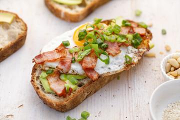Fototapeta na wymiar Open sandwich made of slices of sourdough bread with addition of avocado, egg, bacon, fresh scallion on a wooden white table, close-up. 