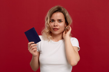 Naklejka premium Portrait of a beautiful girl with curly blond hair dressed in a white t-shirt standing on a red background. Model smiles at the camera and holds passport of blue color.