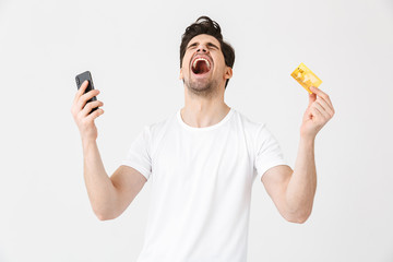Excited happy young man posing isolated over white wall background using mobile phone holding credit card.