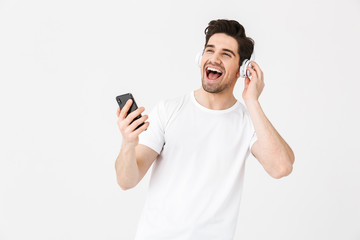 Emotional excited young man posing isolated over white wall background listening music with headphones.