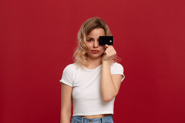 Portrait of a girl with curly blond hair in a white t-shirt standing on a red background. Model looks at the camera and holds bank card covering half of a face.