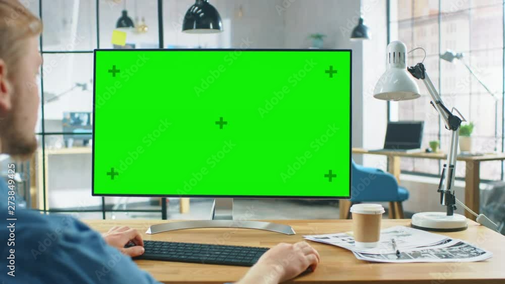 Poster over the shoulder shot: man uses laptop computer with green mock-up screen while sitting at his desk