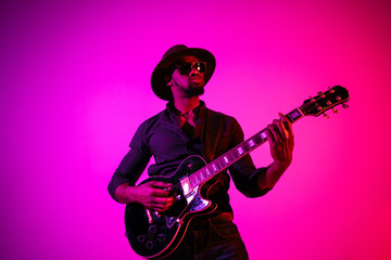 Obraz na płótnie Canvas Young african-american musician playing the guitar like a rockstar on gradient purple-pink background in neon light. Concept of music, hobby. Joyful attractive guy improvising. Colorful portrait.