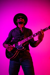 Young african-american musician playing the guitar like a rockstar on gradient purple-pink background in neon light. Concept of music, hobby. Joyful attractive guy improvising. Colorful portrait.