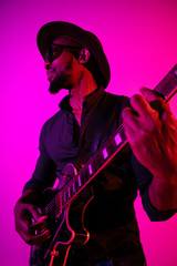 Obraz premium Young african-american musician playing the guitar like a rockstar on gradient purple-pink background in neon light. Concept of music, hobby. Joyful attractive guy improvising. Colorful portrait.
