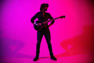Young african-american musician playing the guitar like a rockstar on gradient purple-pink background in neon light. Concept of music, hobby. Joyful attractive guy improvising. Colorful portrait.