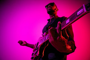 Fototapeta premium Young african-american musician playing the guitar like a rockstar on gradient purple-pink background in neon light. Concept of music, hobby. Joyful attractive guy improvising. Colorful portrait.