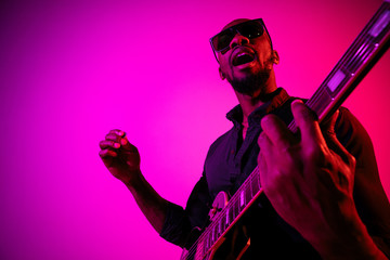 Young african-american musician playing the guitar like a rockstar on gradient purple-pink...
