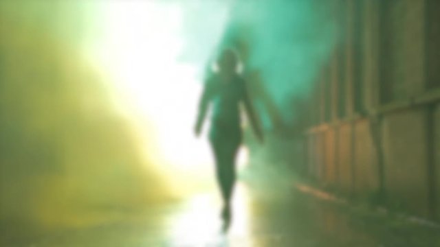 Beautiful Blond woman in leather jacket coming front of the camera in a narrow alley with brick walls with smoke and strobe lightning in the background. Walking on wet tar floor. Spot reflection.