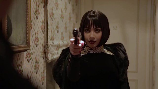 Sexy brunette Arab woman in tight black dress with jewels and big pearl necklace pointing a gun on her husband who cheated on her in a retro hotel with walls adorned with floral tapestry. Slow motion.