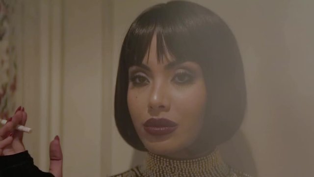 Sexy brunette Arab woman in tight black dress with jewels and big pearl necklace smoking a long cigarette in a retro hotel with walls adorned with floral tapestry. Slow motion. Vengeance.