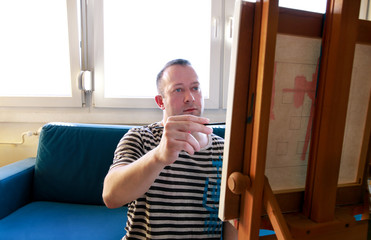 Fototapeta premium Male artist painter working in workshop with canvas on wood drawing board easel in art paint studio. Portrait of artist with paintbrush is painting on canvas with oil paints. Creativity concept.