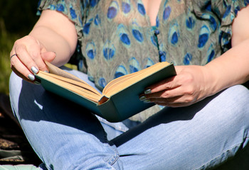 Nice fashion woman lies on green grass and reading an interesting book in city park at summer day. Handsome student woman is sitting and reads book, study and enjoys in beautiful natural environment.