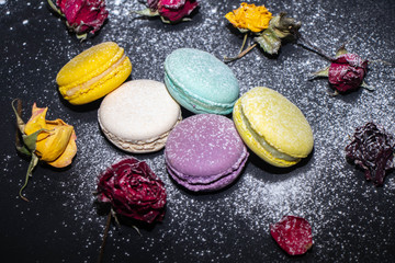 Cakes Macaroons of different colors lie on a dark background, sprinkled with powdered sugar, next to dried flowers of roses. 