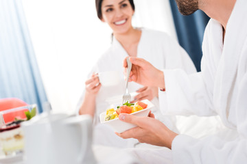 cropped view of man holding fork and bowl with fruit salad near happy woman