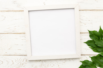 Summer flat lay with white photo frame and green leaves in corner on a white wooden background. Top view mockup with copy space for lettering