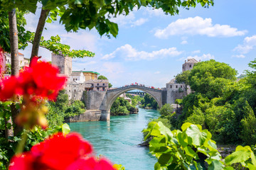A view of the old bridge in Mostar from the terrace through trees, plants and flowers. - obrazy, fototapety, plakaty