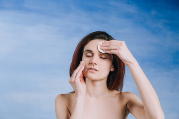 Young woman with a two sponge wihte background cotton pads cleans problem skin with closed eyes on a blue background.
