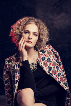 A young male blond androgyn in the image of a beautiful woman sadly sits leaning on her arm, with a summer coat draped over her shoulders with a floral pattern.