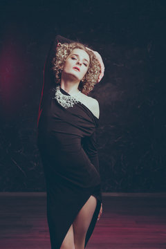 Young male blond androgyn in the image of a beautiful woman sensually posing in a black dress on a dark background